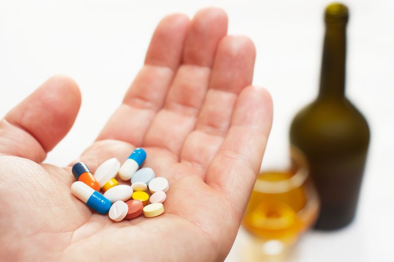 hand holding lots of different coloured and shaped pills with a bottle of alcohol and a glass in the background out of focus