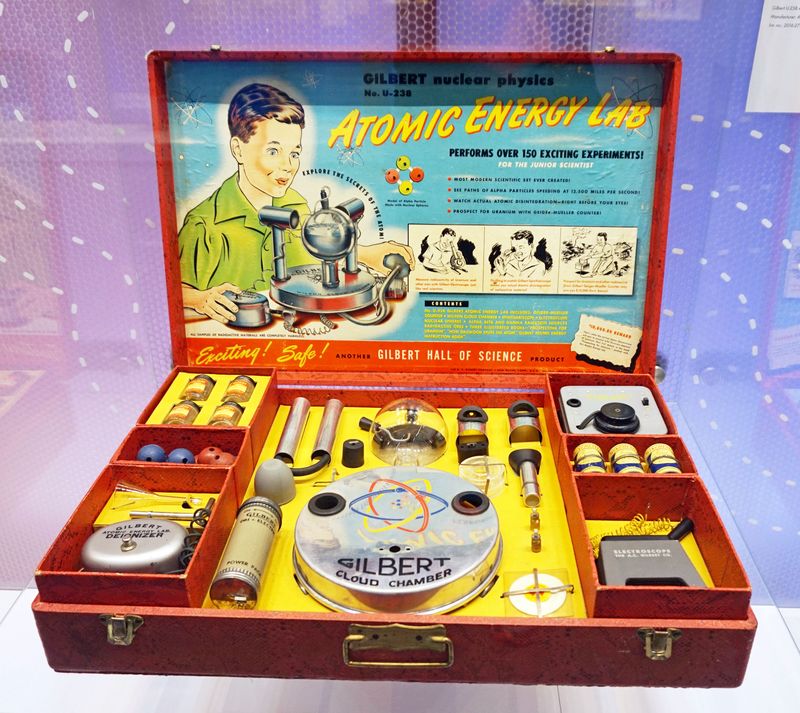The photo shows an example of the Gilbert Atomic Energy Lab in its red box. The box has been separated into several spaces where tools and samples can be stored. The lid of the box includes an information sheet fitted into it that shows a cartoon boy looking at the assembled pieces, as well as instructions for its use.  