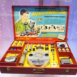 The photo shows an example of the Gilbert Atomic Energy Lab in its red box. The box has been separated into several spaces where tools and samples can be stored. The lid of the box includes an information sheet fitted into it that shows a cartoon boy looking at the assembled pieces, as well as instructions for its use.  
