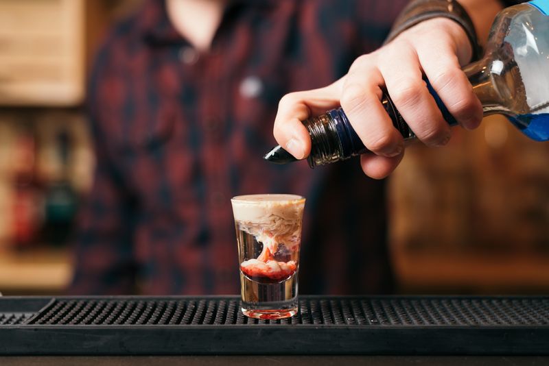 Handsome bearded barman is making monkey brain cocktail in night club. Selective focus on glass.