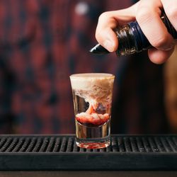 Handsome bearded barman is making monkey brain cocktail in night club. Selective focus on glass.