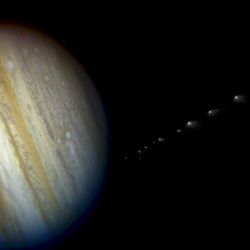 Jupiter is seen on its side with Io casting a small shadow on it. On the side small blurry fragements are seen trailing, the comet fragments approaching the planets