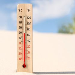 wooden thermometer with red scale showing celsius and fahrenheit, sitting in white sand on a beach