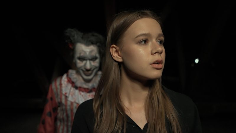 Evil-looking sinister clown in a white makeup face and red and white stripy clothes in a blurred background behind a caucasian female who is looking worried. 