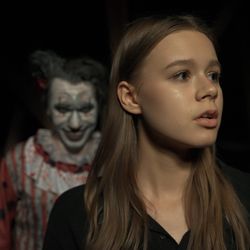 Evil-looking sinister clown in a white makeup face and red and white stripy clothes in a blurred background behind a caucasian female who is looking worried. 