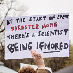 A protestor holds up a cardboard signs saying "At the start of every disaster movie there's a scientist being ignored". The words "disaster movie" are written in red, while the rest of the text is written in black. The photo puts the sign in the centre of the image, with the backs of a few people's heads in the foreground. In the background, a leafless tree is visible suggesting the photo was taken in winter. 