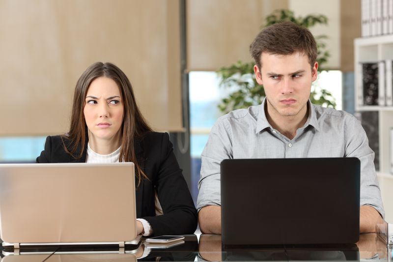 A woman and a man sit next to one another in an office space. They both have their laptops in front of them at the shared table. The woman is wearing a black blazer and a white jumper while the man is wearing a stone-grey shirt. The two are side eyeing one another and looking irritated. 