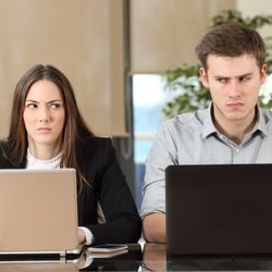 A woman and a man sit next to one another in an office space. They both have their laptops in front of them at the shared table. The woman is wearing a black blazer and a white jumper while the man is wearing a stone-grey shirt. The two are side eyeing one another and looking irritated. 
