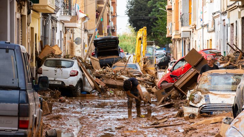 A road in the city is covered in debris and mud, not just on the pavement but also on cars. One person is pouring a bucket of mud on the streets