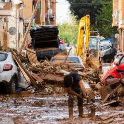 A road in the city is covered in debris and mud, not just on the pavement but also on cars. One person is pouring a bucket of mud on the streets