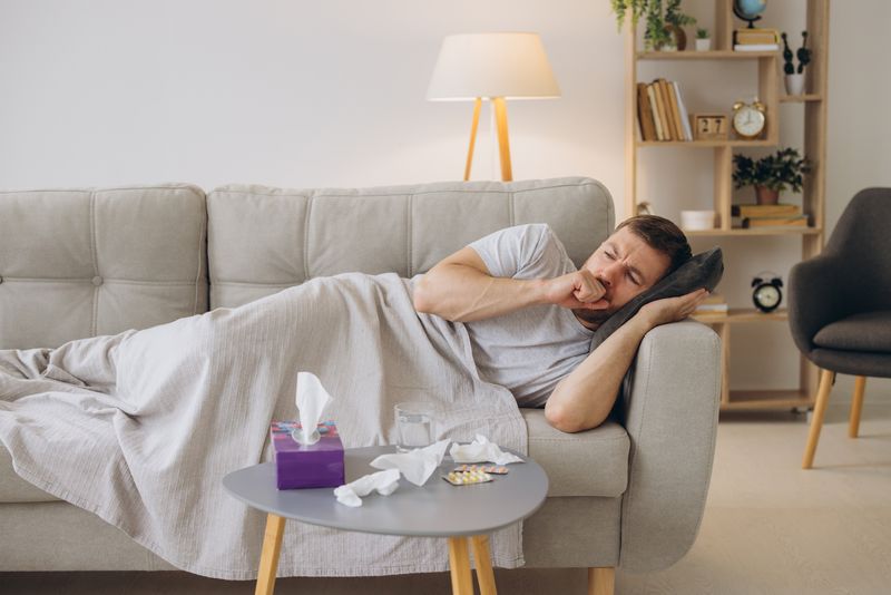 man lying on the sofa under a blanket, coughing; there are tissues on the table in front of him