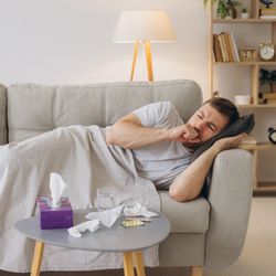 man lying on the sofa under a blanket, coughing; there are tissues on the table in front of him