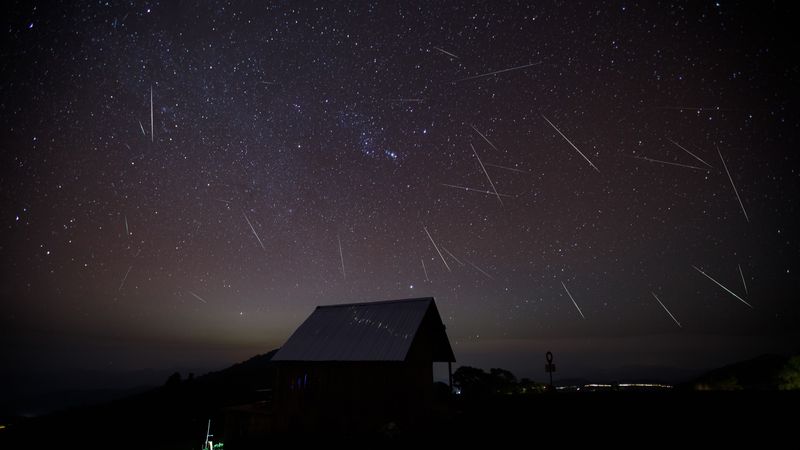 A hut is seen in the foreground and behind the night sky is streaked by meteors