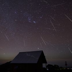A hut is seen in the foreground and behind the night sky is streaked by meteors