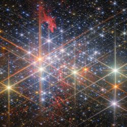 A dense cluster of bright stars, each with six large and two small diffraction spikes, due to the telescope’s optics. They have a variety of sizes depending on their brightness and distance from us in the cluster, and different colours reflecting different types of star. Patches of billowing red gas can be seen in and around the cluster, lit up by the stars. Small stars in the cluster blend into a background of distant stars and galaxies on black.]