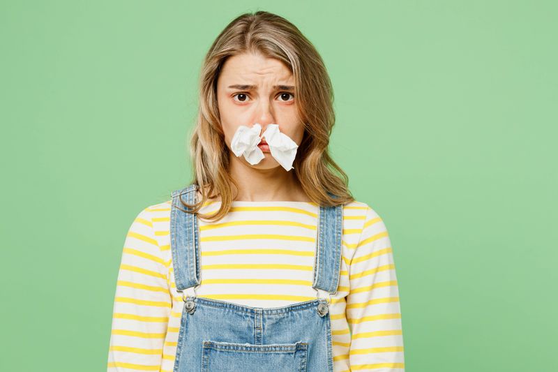 A woman wearing a yellow and white stripy long-sleeve t-shirt and dungarees is looking at the camera. She has two tissues stuffed up her nose and se looks sorry for her self. She is set against a pea green background.  