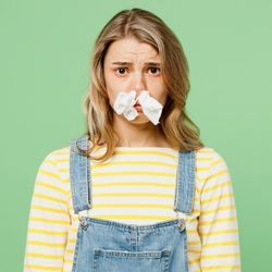 A woman wearing a yellow and white stripy long-sleeve t-shirt and dungarees is looking at the camera. She has two tissues stuffed up her nose and se looks sorry for her self. She is set against a pea green background.  