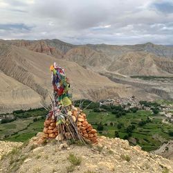 A photo showing a monument made of piled clay-red stones and thin sticks assembled around what looks like different colored flags. The monument is on a hillside looking down on a Tibetan village nestled within green fields among harsh, barren looking hillsides. 