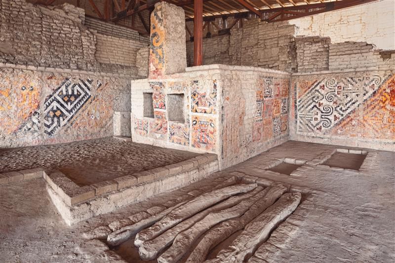 The photo shows the interior of Huaca Cao Viejo. It is an open space that is walled off. In the far corner, there is a smaller square structure with two smaller square spaces in it, like windows. This structure and the surrounding walls are covered in painted frescos showing intricate designs. 