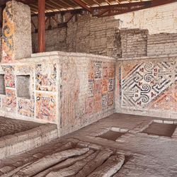 The photo shows the interior of Huaca Cao Viejo. It is an open space that is walled off. In the far corner, there is a smaller square structure with two smaller square spaces in it, like windows. This structure and the surrounding walls are covered in painted frescos showing intricate designs.