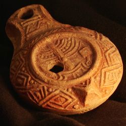 The photo shows the lamp lit by a soft orange light. The lamp is roughly tear-drop shaped and made of ceramic. It's surface has intricate decorations across it and, in the center, is the symbol of the menorah.