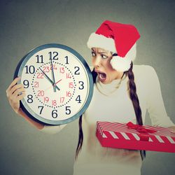 A woman wearing a Santa's hat is holding a clock with the time (11:00) and the dates on it (23rd December). She looks shocked and worried. She is wearing a white jumper and holding a red and white striped wrapped box in the other hand.
