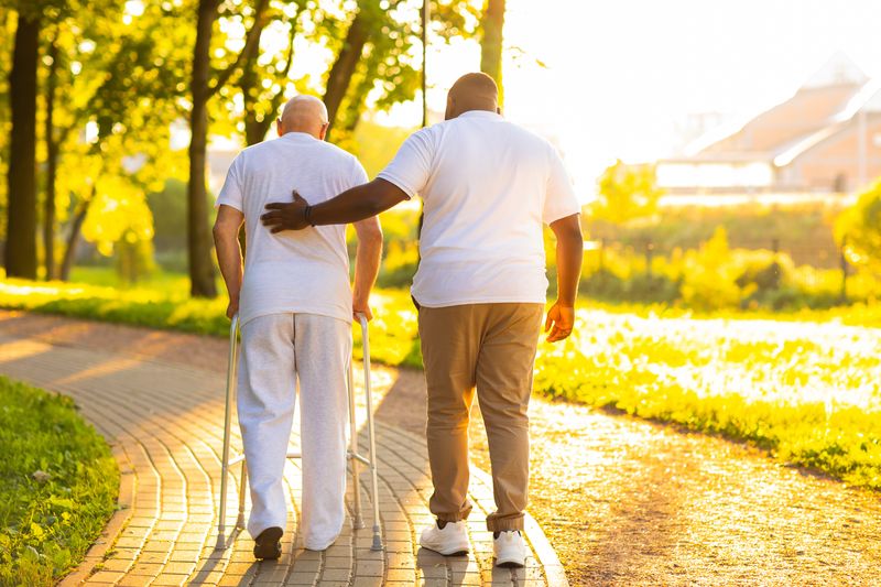 man wearing white walking with a zimmer frame while another man guides him with a hand on his back; they are viewed from behind, walking down a paved path next to some grass and trees
