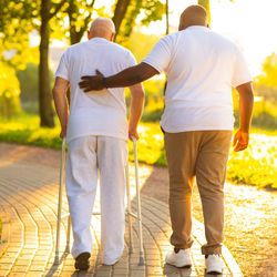 man wearing white walking with a zimmer frame while another man guides him with a hand on his back; they are viewed from behind, walking down a paved path next to some grass and trees