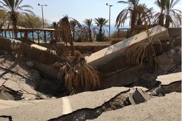 A sinkhole in the middle of a road. The ground has collapsed in and the debris includes what appear to be a bus and a lory. There are palm trees growing in the middle of fissure. In the background, the lake is visible against a blue sky. A sinkhole in the middle of a road. The ground has collapsed in and the debris includes what appear to be a bus and a lory. There are palm trees growing in the middle of fissure. In the background, the lake is visible against a blue sky.