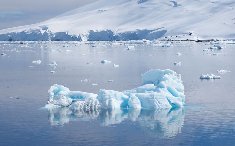 The photo shows a piece of ice floating in a still body of water with a snow covered mountain behind it. The ice has a bluish tinge to it and there are other pieces of ice scattered across the water.