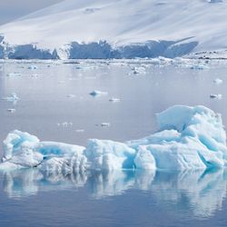 The photo shows a piece of ice floating in a still body of water with a snow covered mountain behind it. The ice has a bluish tinge to it and there are other pieces of ice scattered across the water.