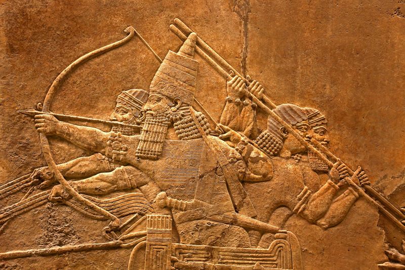 An ancient Assyrian relief showing a soldiers in a chariot holding spears and bows as they rise into battle.  