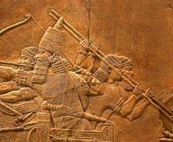 An ancient Assyrian relief showing a soldiers in a chariot holding spears and bows as they rise into battle.  