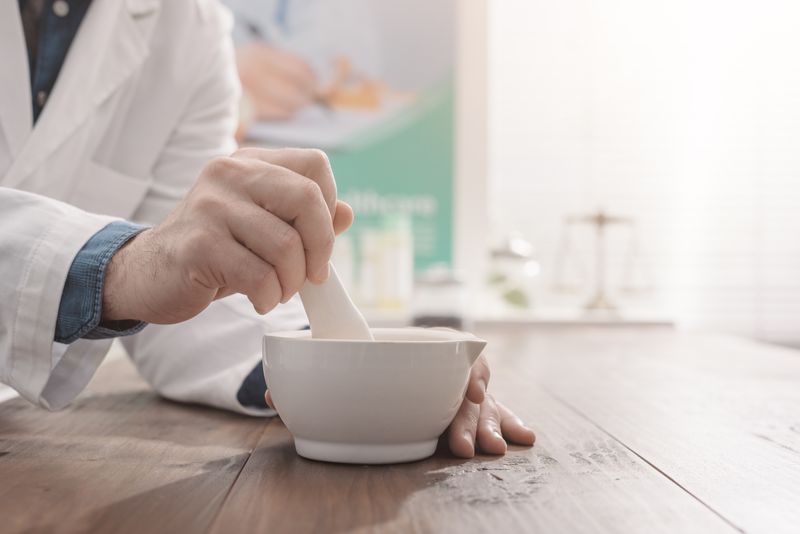 close up of someone's hands using a mortar and pestle; they are wearing a white labcoat over a blue shirt and standing at a wooden bench, with a set of scales out of focus in the background