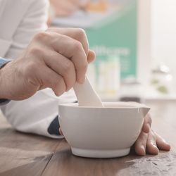 close up of someone's hands using a mortar and pestle; they are wearing a white labcoat over a blue shirt and standing at a wooden bench, with a set of scales out of focus in the background