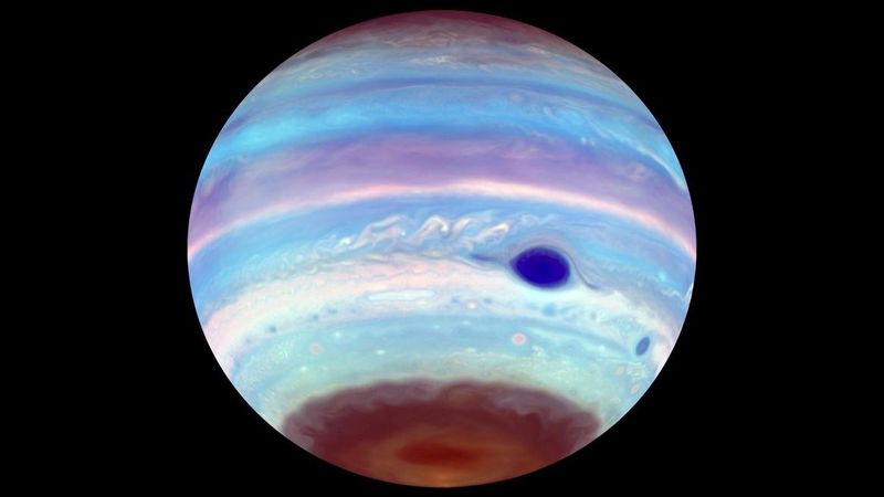 The band and clouds of jupiter are visible in this image. the great red spot is starkingly clear in a dark color. The souther pole looks dimmer covered in this haze and among the haze a dark patch where the oval is