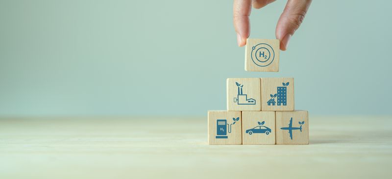 Photo of building block pyramid with various things that consume fuel on, like a car and a plane, with a hand placing a hydrogen block on top
