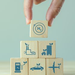 Photo of building block pyramid with various things that consume fuel on, like a car and a plane, with a hand placing a hydrogen block on top