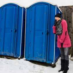 woman wearing a pink coat and black hat, scarf and gloves waiting by two blue portapotties outside in the snow