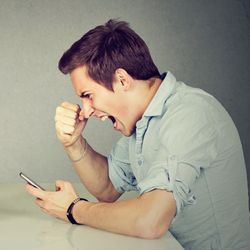 man and woman sitting opposite each other on a table looking angrily at a phone and laptop respectively