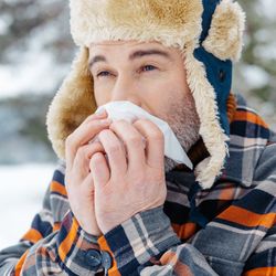 A man in a checkered coat and a fake-fur lined trapper hat is blowing his nose. He is standing in what looks like a snow covered field with a tree in the background. 