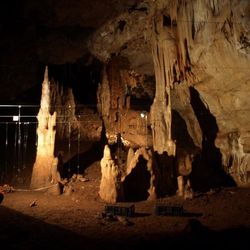 The photo shows the large cavern in the Manot Cave. It has been illuminated by artificial light revealing the cave wall with several rock features sticking up from the ground. The image shows a researcher to the right kneeling and looking away from the camera as they examine a lattice of cables that have been set up as a kind of frame. 