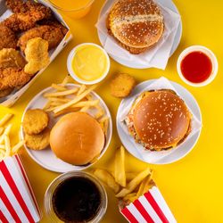 The top down look at food on a yellow surface. The food has been gathered to the left of the shot and shows three burgers - one with a plain bun - surrounded by fires and chicken nuggets. There are also some ramekins with different colored dips in them. The fries are served in red and white striped boxes.