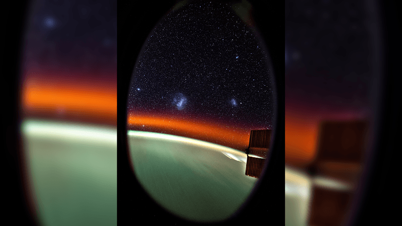 The view from the cupola of the ISS showing Earth as a light blurry green, an orange band of airglow and two small galaxies, which are the small and large Magellanic clouds. 