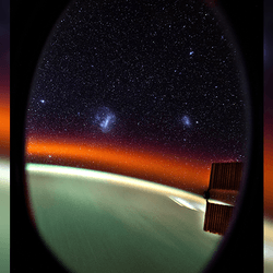 The view from the cupola of the ISS showing Earth as a light blurry green, an orange band of airglow and two small galaxies, which are the small and large Magellanic clouds. 