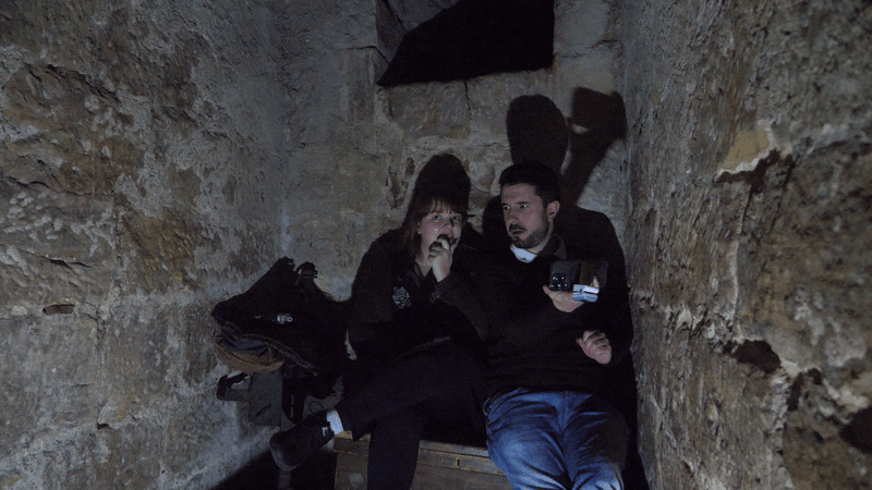Two scared looking people (male and female) sitting in Chillingham's haunted dungeon. Both are looking scared. 