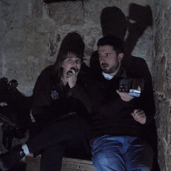 Two scared looking people (male and female) sitting in Chillingham's haunted dungeon. Both are looking scared. 