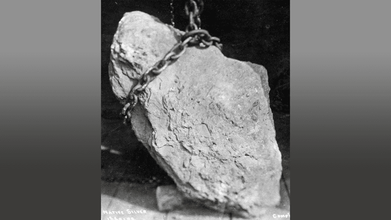 A large silver nugget in a black and white photo. A big chain is around the top portion of the nugget.