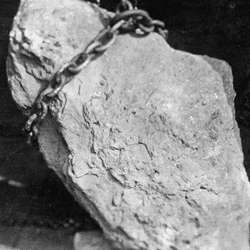 A large silver nugget in a black and white photo. A big chain is around the top portion of the nugget.